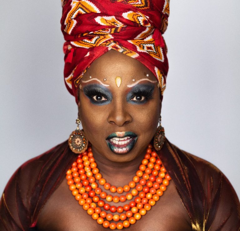 A black drag queen looks directly at the camera. She is wearing a bright red and orange headscarf, has vibrant makeup and a large orange bea