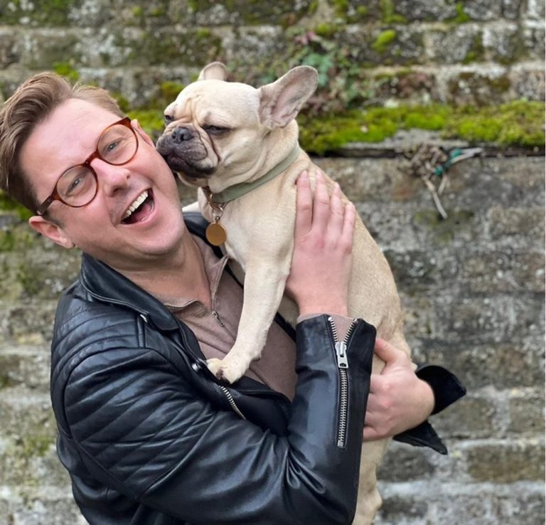 A blonde man laughing at the camera in front of a brick wall. He's wearing a black leather jacket and holding a grumpy-looking fawn French b
