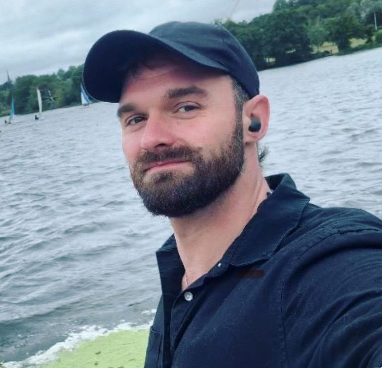 A selfie of a man smiling at the camera. He has a beard and wearing a dark cap and navy polo shirt. Behind him is a lake with sail boats.
